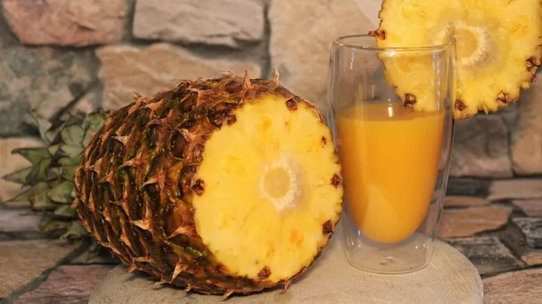 Poloviční ananas vedle sklenice čerstvého džusu na kamenném povrchu.