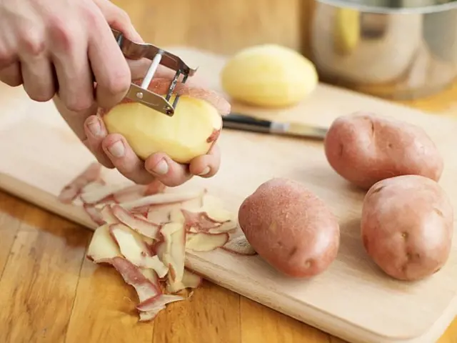 Člověk škrábe kůru z brambor na dřevěném prkénku.