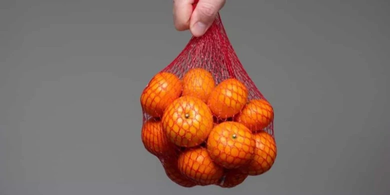 Ruka drží sáček plný oranžových citrusů na šedém pozadí.